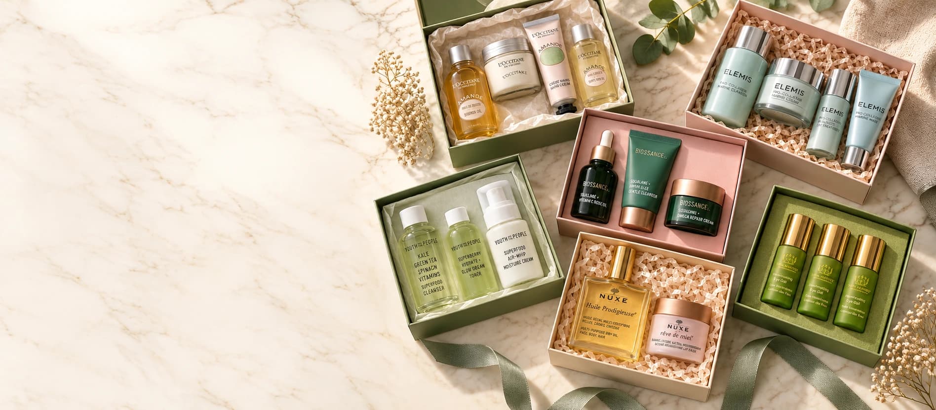 An elegant flat-lay of premium beauty gift sets with travel-size glass bottles in gift boxes on a cream marble surface.